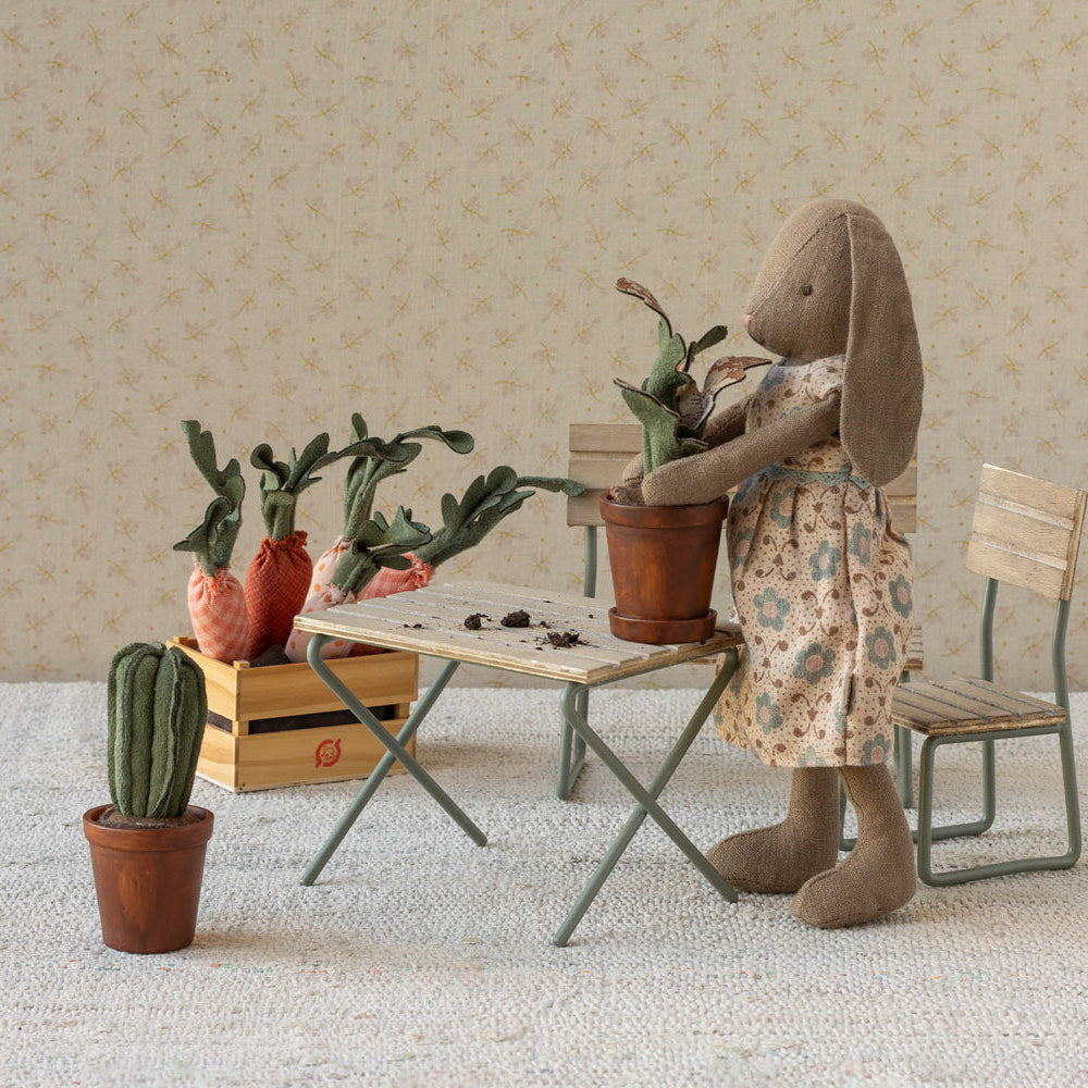 A Maileg toy rabbit wearing a floral dress stands by a miniature table and chairs, holding a small potted plant. Other miniature potted plants, carrots in a crate, and a cactus sit nearby in a cozy room setting with wallpaper.