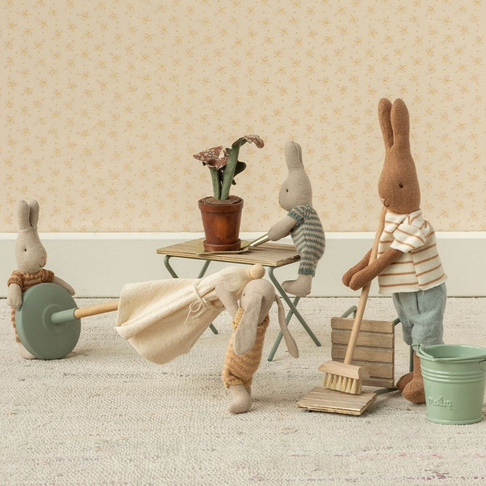 Three Maileg toy rabbits are gathered in a miniature room with a table, chairs, a broom, a bucket, and a crate of carrots, creating a playful scene against patterned wallpaper.