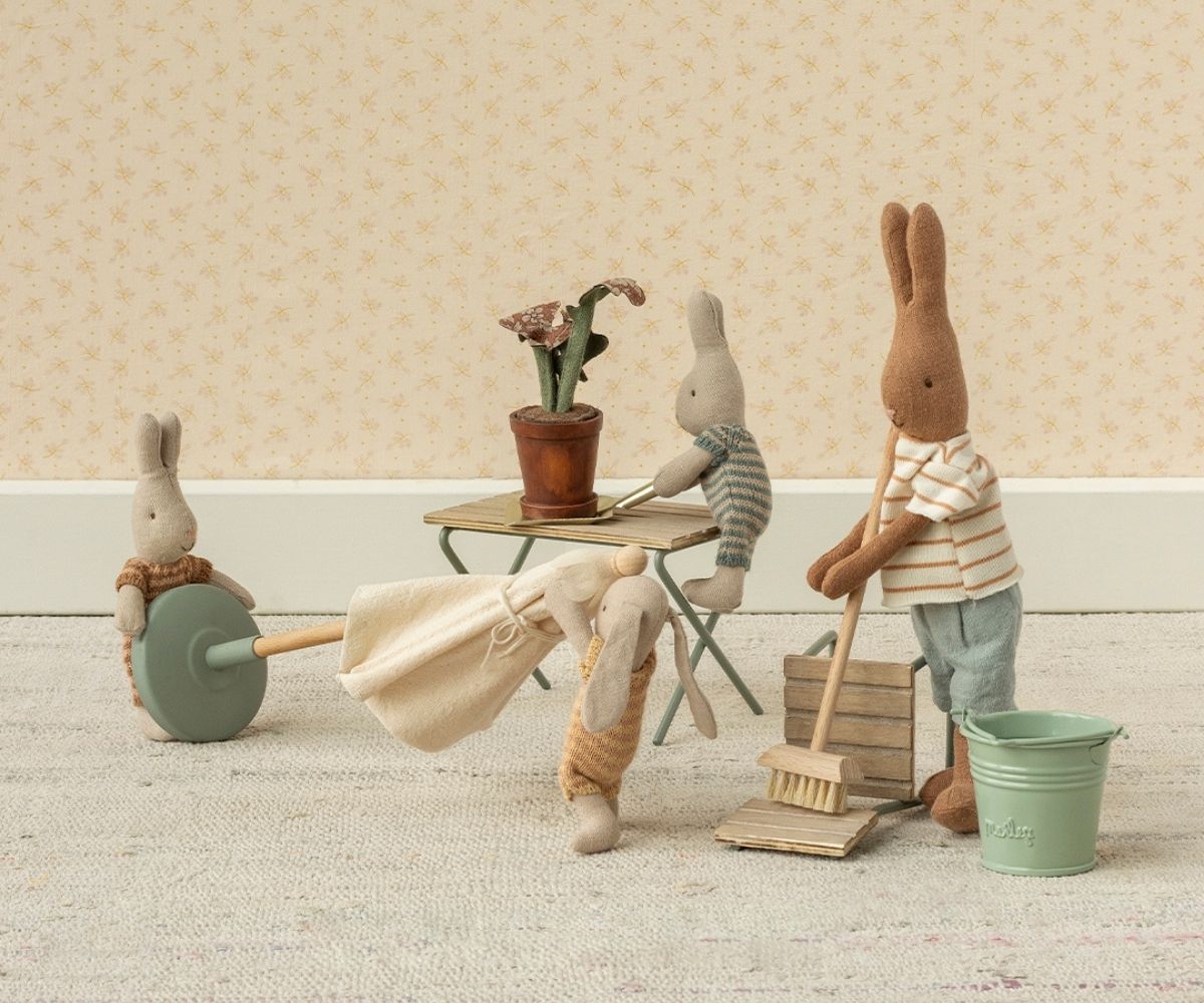 Three Maileg toy rabbits are gathered in a miniature room with a table, chairs, a broom, a bucket, and a crate of carrots, creating a playful scene against patterned wallpaper.
