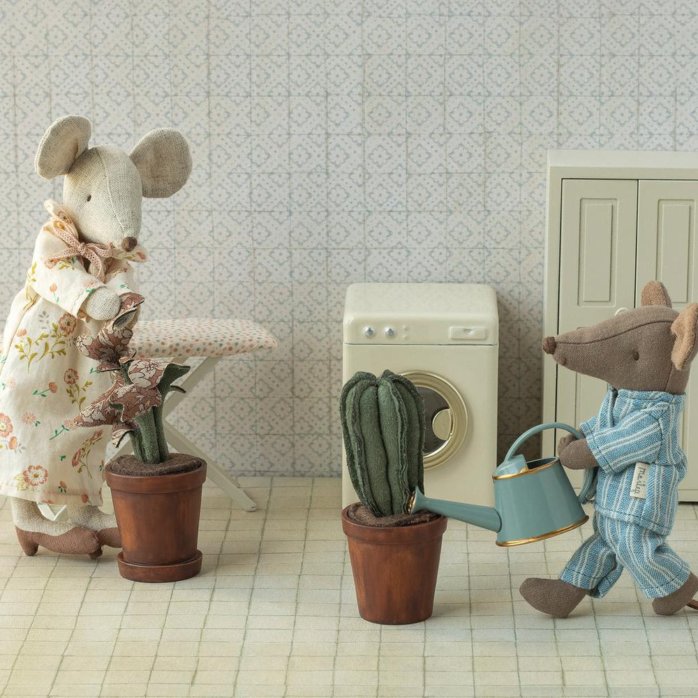 A Maileg toy mouse in striped pajamas holding a blue watering can walks toward a potted cactus, while another Maileg mouse in a floral dress stands by a table holding a small potted plant in a miniature room with patterned wallpaper.