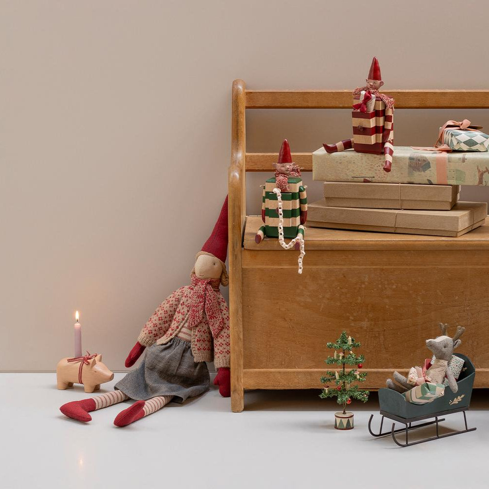 A miniature green wooden Maileg toy mouse sleigh on a wooden floor, with a larger plush toy duck, gnome dolls, a small suitcase, and holiday decor arranged in a rustic room setting.
