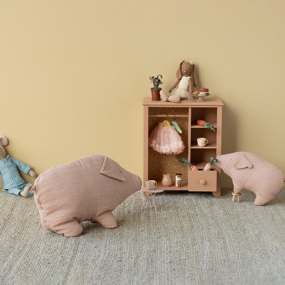A soft, dusty pink linen stuffed pig toy standing next to a miniature wooden wardrobe on a textured rug, alongside a plush mouse and a larger pillow-like pig.