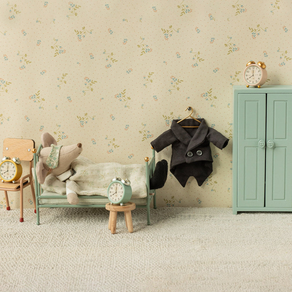 A Maileg mouse toy relaxing in a mint green miniature metal single bed next to a small wooden chair with a clock, a hanging jacket on a hook, a mint green storage cabinet with a clock on top, and floral wallpaper in the background.