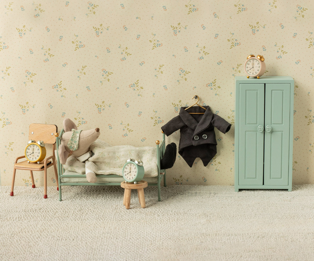 A Maileg mouse toy relaxing in a mint green miniature metal single bed next to a small wooden chair with a clock, a hanging jacket on a hook, a mint green storage cabinet with a clock on top, and floral wallpaper in the background.