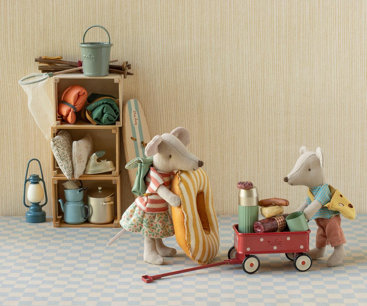 A styled indoor scene with two plush mouse figures dressed in picnic clothing, one carrying a striped donut and the other pushing a red wagon loaded with picnic items, beside a wooden shelf with camping accessories and a lantern.