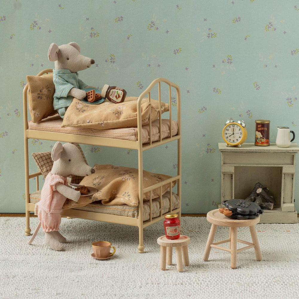 A miniature scene featuring two Maileg mice in a cozy room with floral wallpaper and a bunk bed, where one mouse sits on the top bunk and the other stands near a small wooden stool set up with the Maileg Waffle Set, including a waffle maker, cups, and jars.