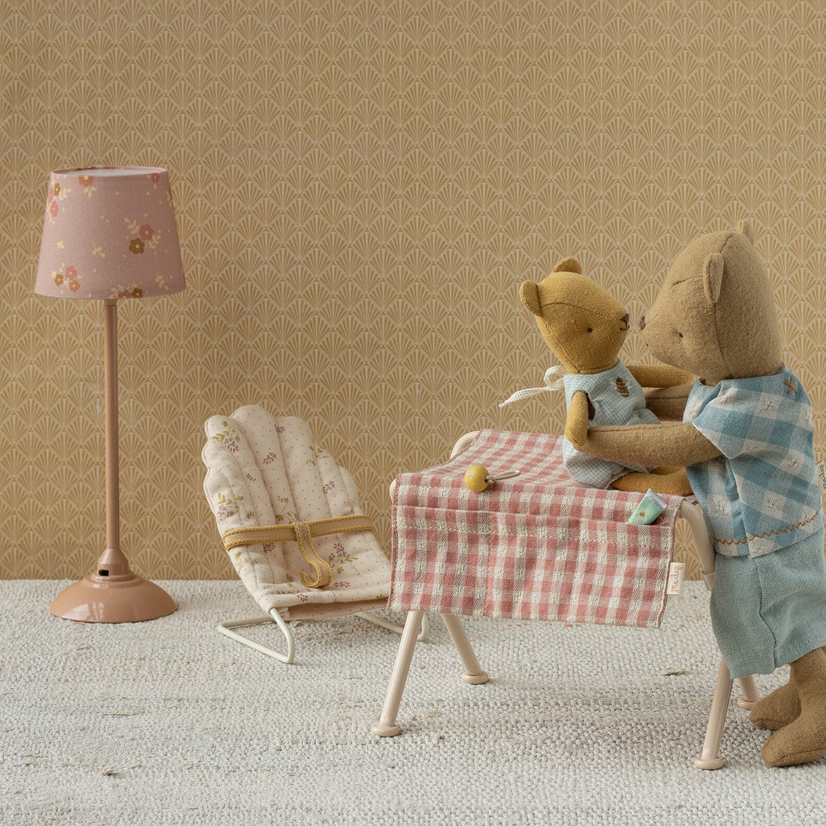 A miniature Maileg nursery scene with a floor lamp, a bouncy chair, and a small table covered in a pink gingham cloth where two toy teddy bears are seated, set against a warm beige patterned wall.