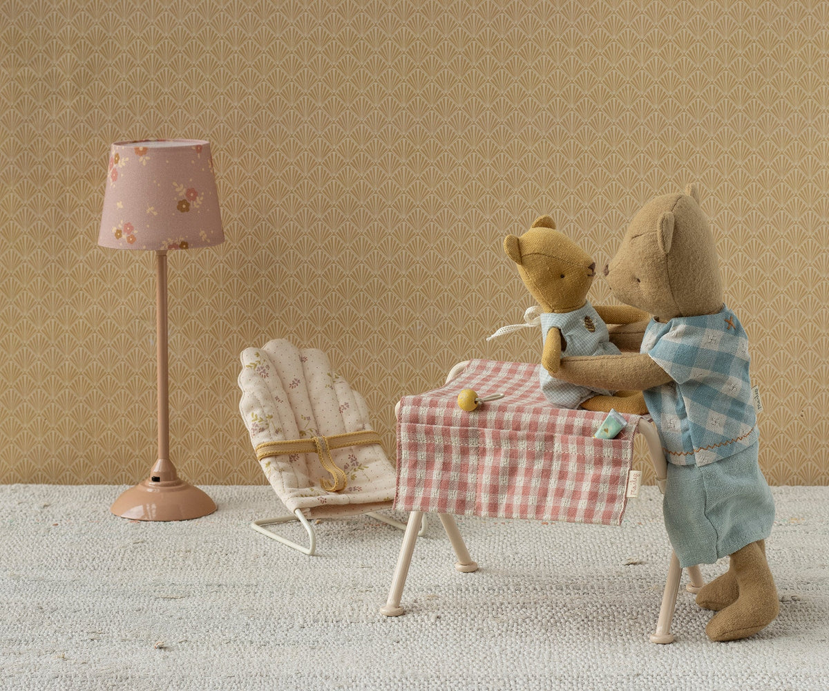 A miniature Maileg nursery scene with a floor lamp, a bouncy chair, and a small table covered in a pink gingham cloth where two toy teddy bears are seated, set against a warm beige patterned wall.