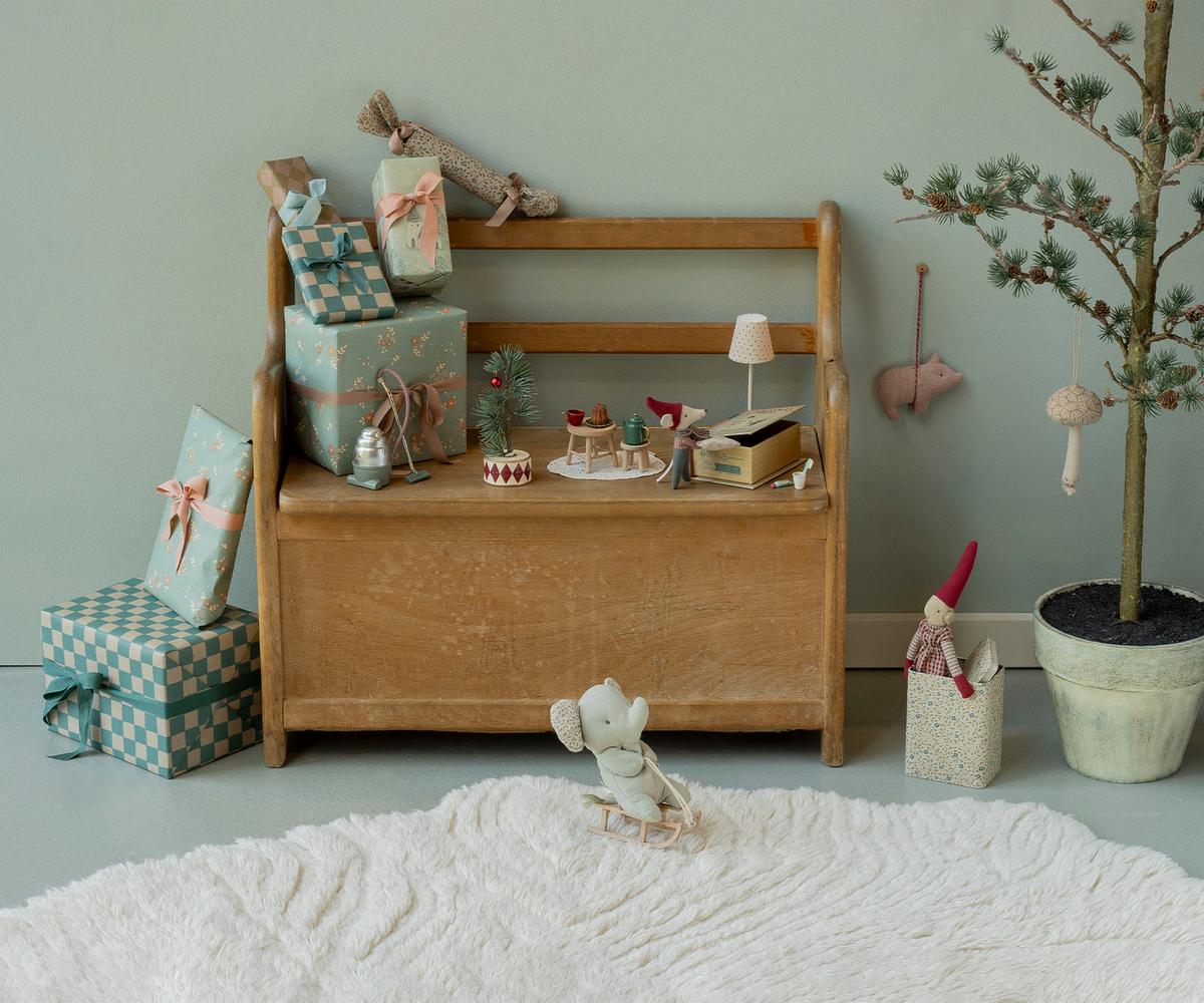 A wooden toy bench styled with wrapped gift boxes, small decorative items, and a plush mouse in a cozy nursery setting against a sage green wall.