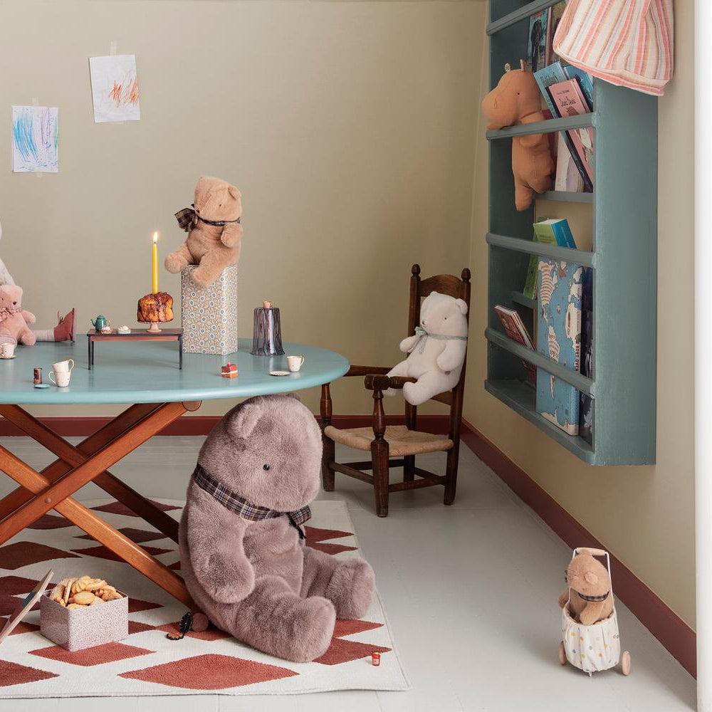 A cozy children's playroom scene featuring a large plush teddy bear wearing a plaid bow sitting on a patterned rug in the foreground, with a teal table, toy chairs, and various stuffed animals and toys in the background.