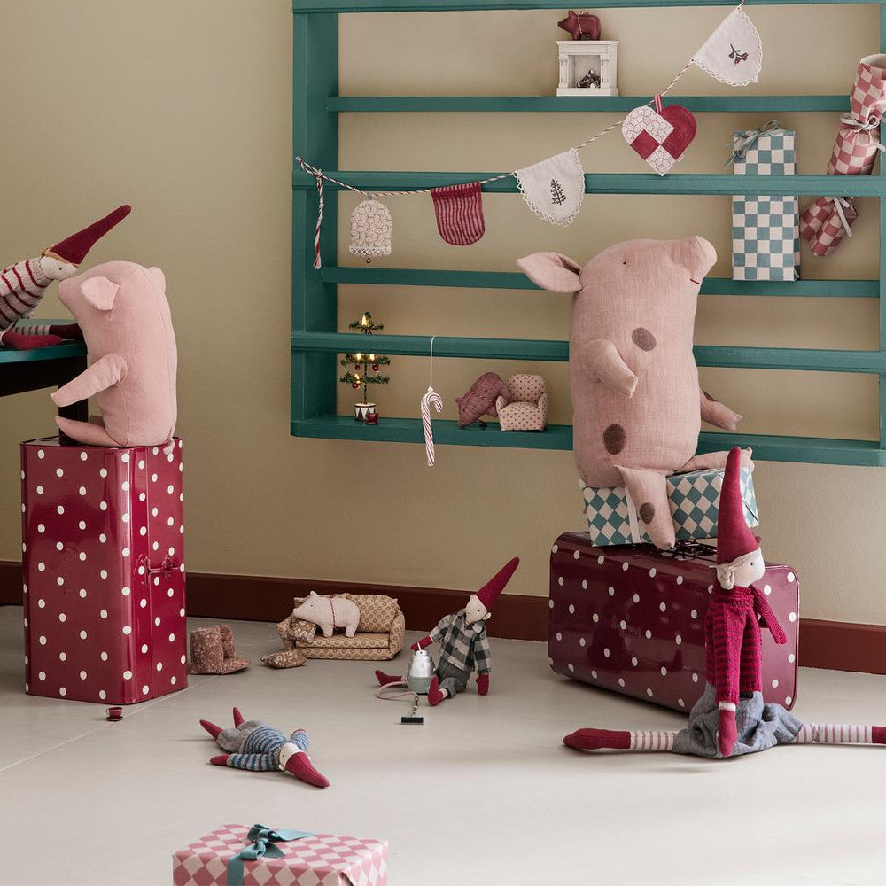 A toy room scene featuring Maileg mice characters: one sitting on a tall burgundy gift box at a small wooden table, another leaning against a large matching suitcase, and a toy dog lying near a miniature sofa bed against a teal wooden wall rack decorated with bunting.