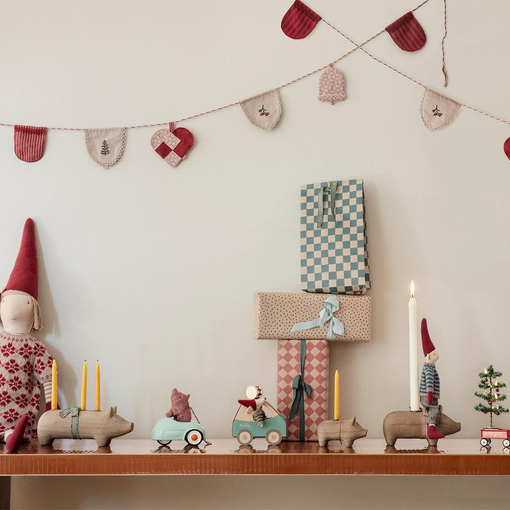 A festive holiday display featuring a mantelpiece decorated with wooden animal figures, miniature toy mice in toy cars, a lit candle, and wrapped gift boxes against a white wall strung with a felt heart and stocking garland.