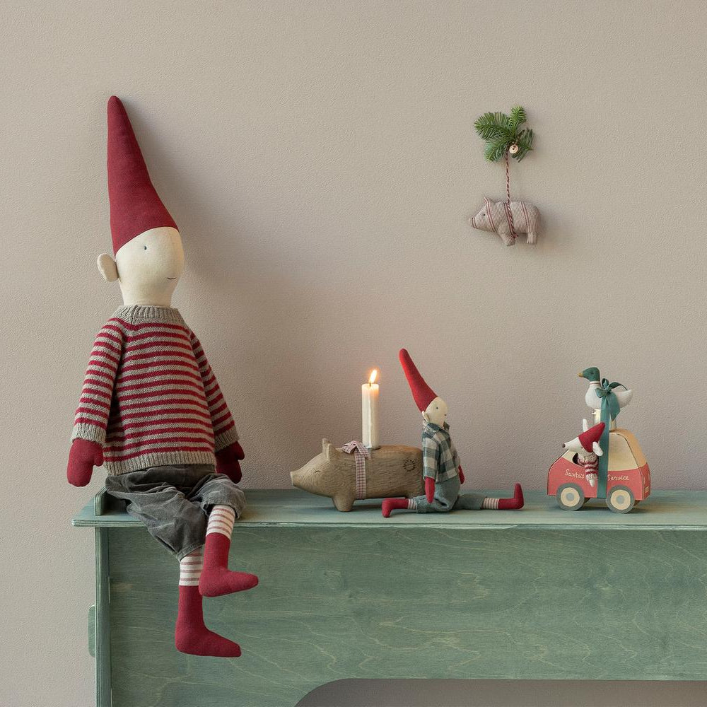 A festive holiday display featuring a large Maileg mouse doll in a red and white striped sweater and a tall pixy hat seated on a mint green ledge, with smaller Maileg mice, a miniature car-shaped box, and a toy pig figurine arranged nearby in front of a warm-toned background.