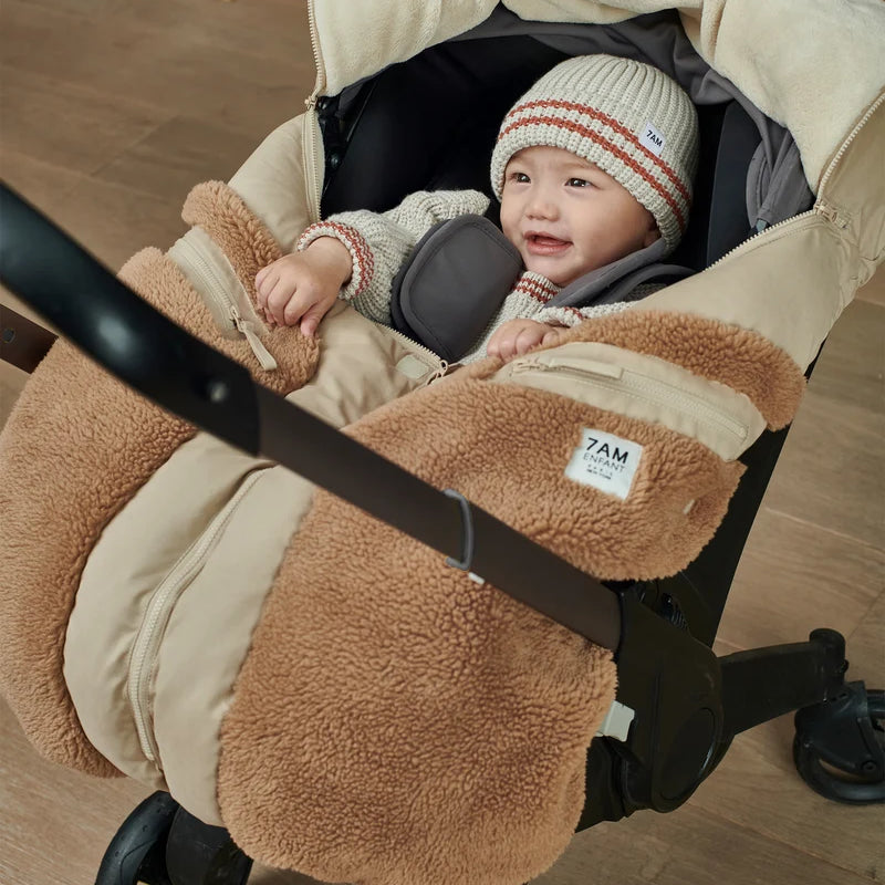 A baby wearing a striped beanie and pacifier is cozy inside an infant car seat outfitted with the Benji Applique Car Seat Cocoon by 7AM Enfant in oat with brown sherpa fleece trim.