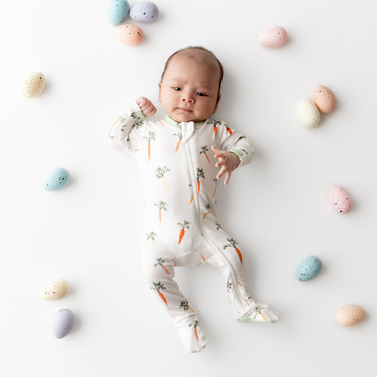 Baby in a white onesie with carrot pattern surrounded by colorful eggs on a light background