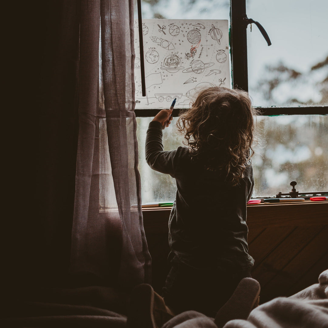 Child drawing on a window with a pen, sitting by a window with a view of trees.