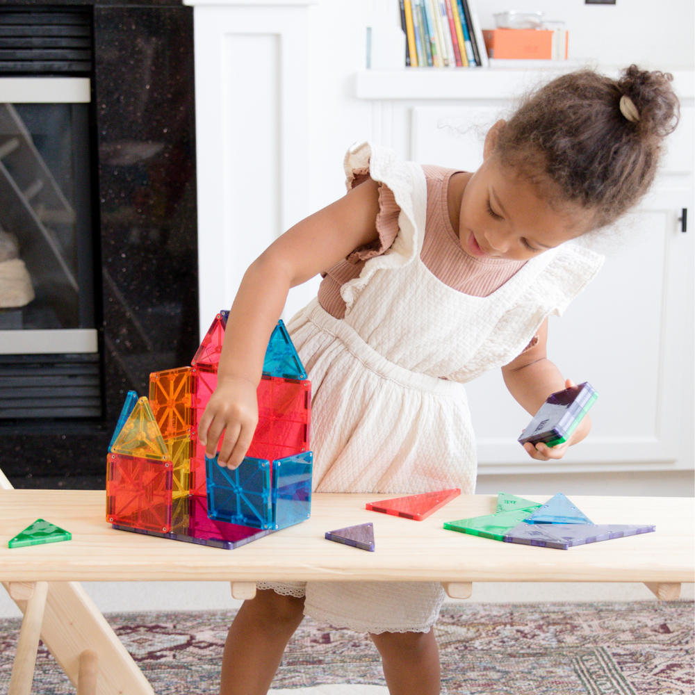 A young child playing on a wooden floor, carefully placing colorful, translucent Magna-Tiles into a structure on a low table.