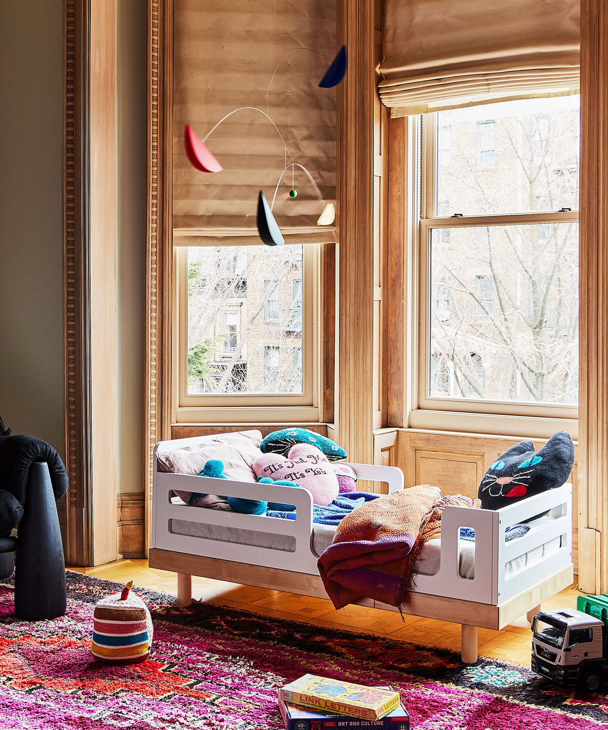 Children's room with a crib, colorful rug, and toys near large windows.