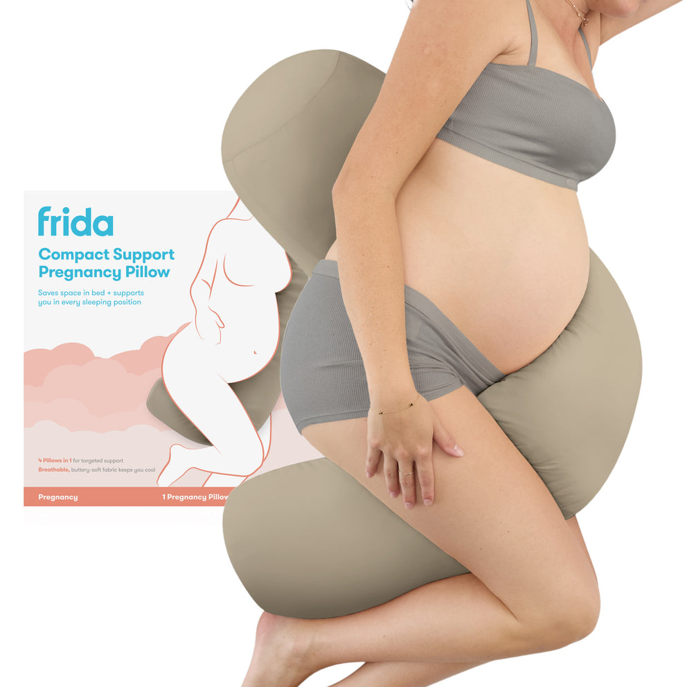 A pregnant model wearing gray maternity underwear sits on a beige compact support pregnancy pillow, with the Frida product box shown to the left on a white background.