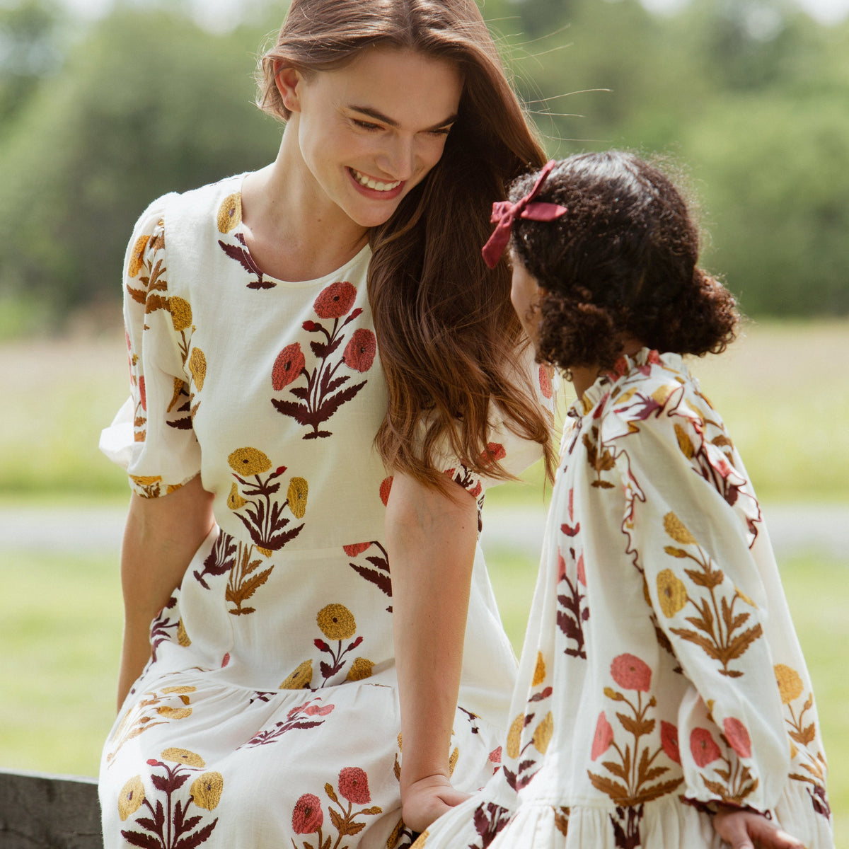 Mother and daughter sitting together outdoors; the mother smiles down at the young girl, both wearing matching cream long-sleeve dresses featuring a golden and coral harvest dandelion print.