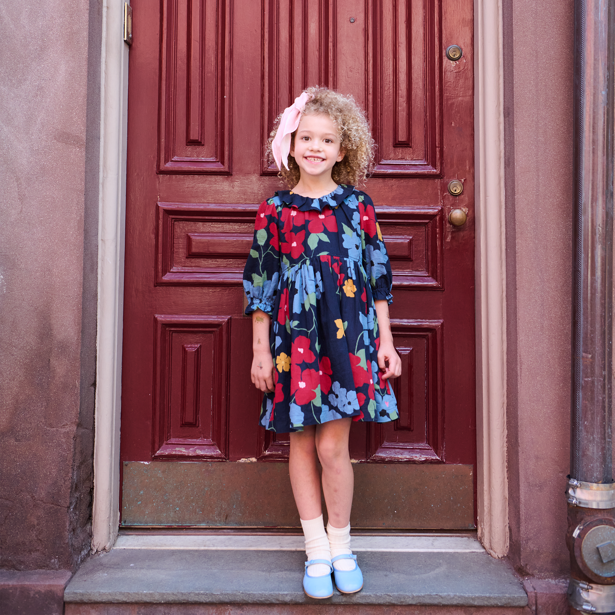 A young girl with curly blonde hair stands on a doorstep in front of a dark red wooden door, wearing a navy blue floral dress with red, yellow, and light blue flowers, white socks, and light blue shoes.