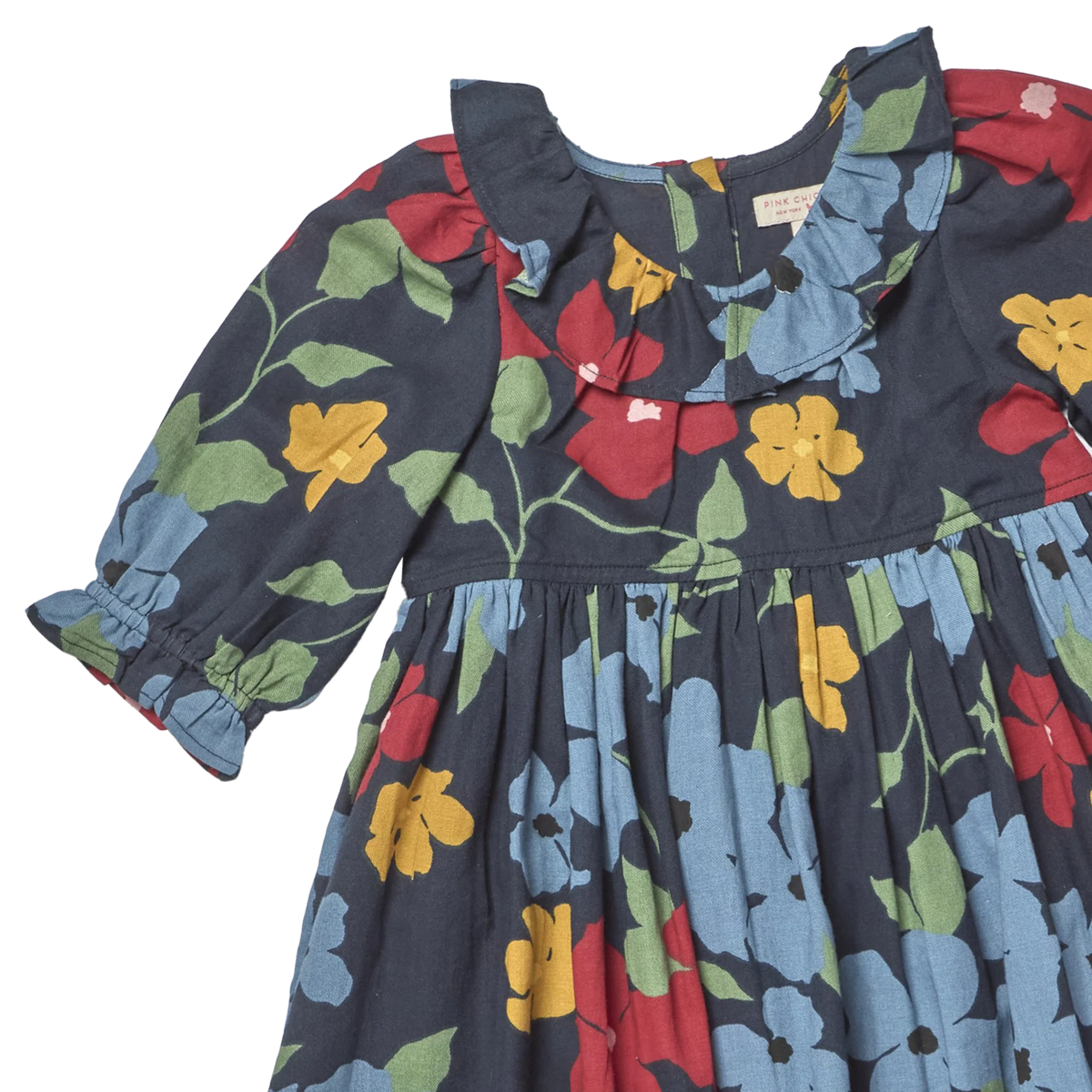 A close-up of a children's dress with a dark navy blue base and a colorful floral print featuring red, yellow, and light blue flowers with green leaves. The dress has a ruffled collar, long sleeves with elastic cuffs, and a gathered waist.