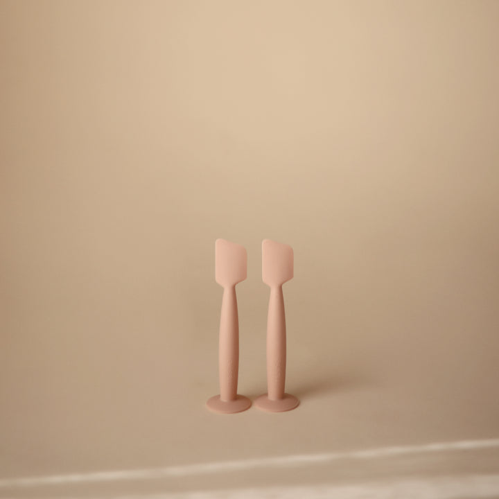Two pale pink silicone spatulas standing upright on a matching round base against a warm beige background.