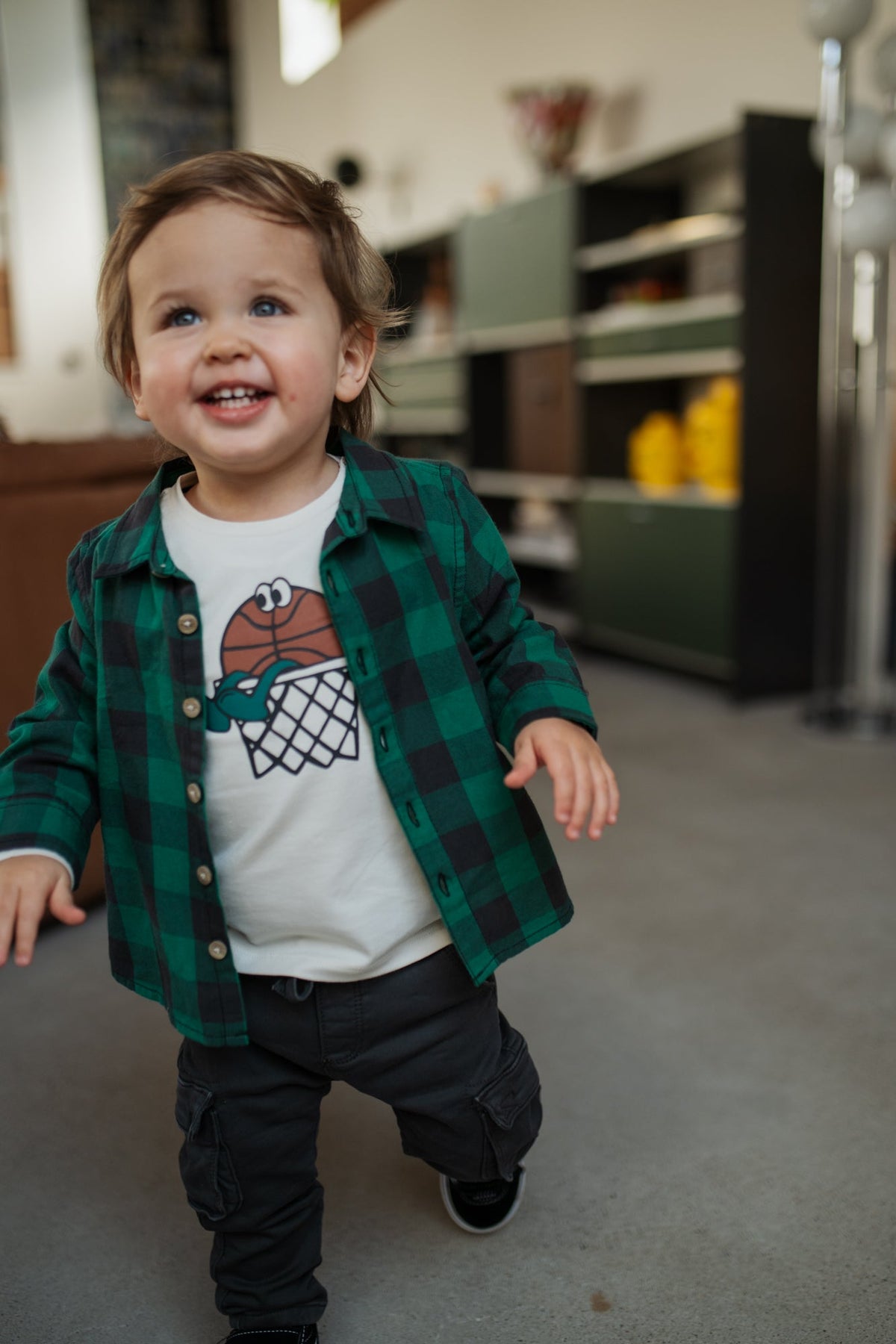 Child wearing a green plaid shirt over a white t-shirt with a graphic design, standing in a room.