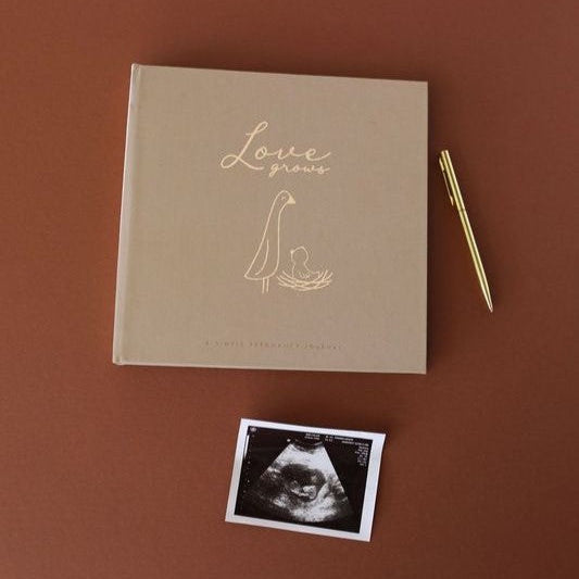 A square tan keepsake book with gold foil lettering reading 'Love grows' and a simple baby silhouette, displayed on a warm brown background with a gold pen and an ultrasound photo print below it.