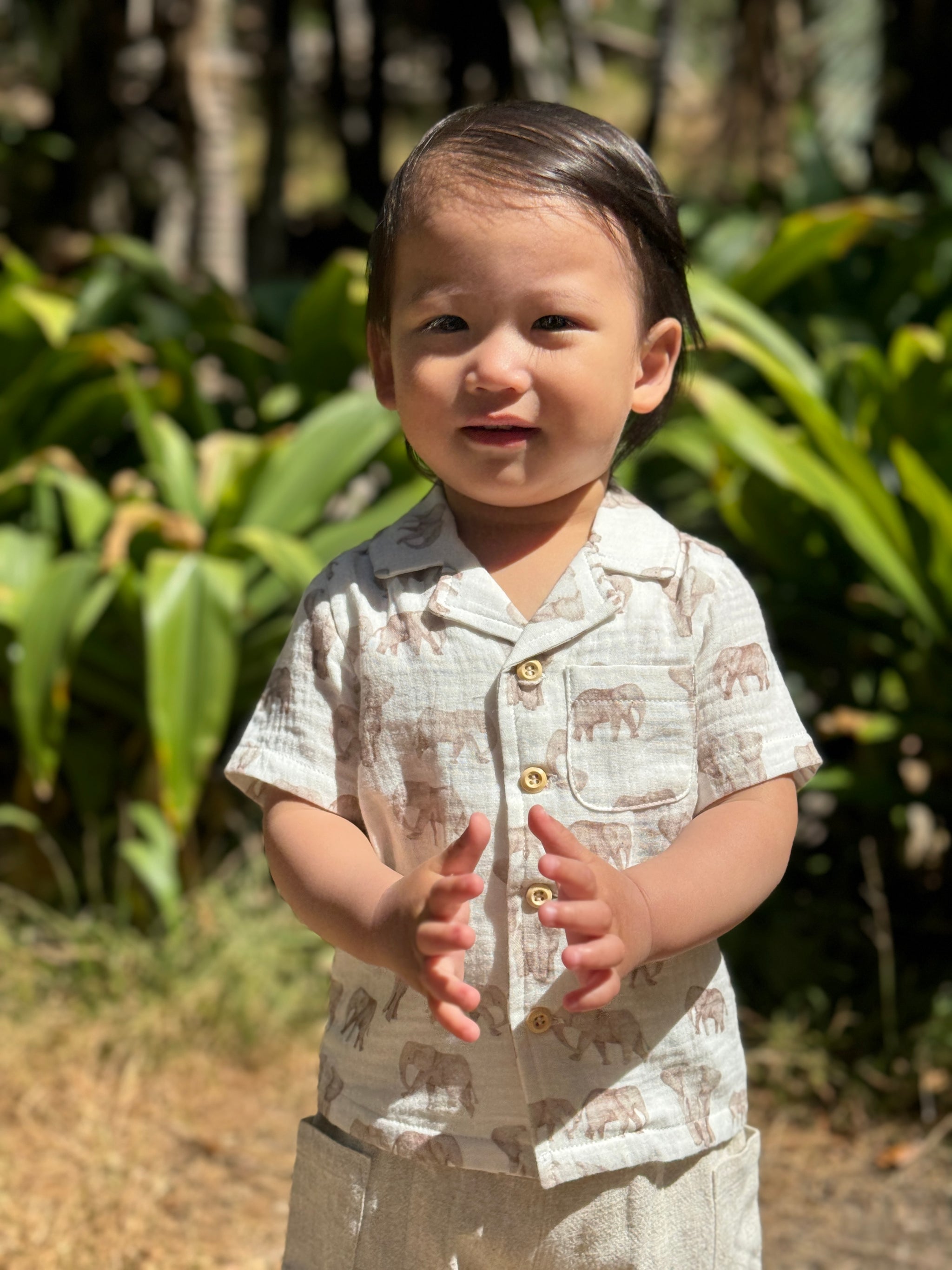 A toddler standing outdoors in front of lush green foliage, wearing a short-sleeve button-front shirt and matching pants in a light beige fabric with a brown animal print.