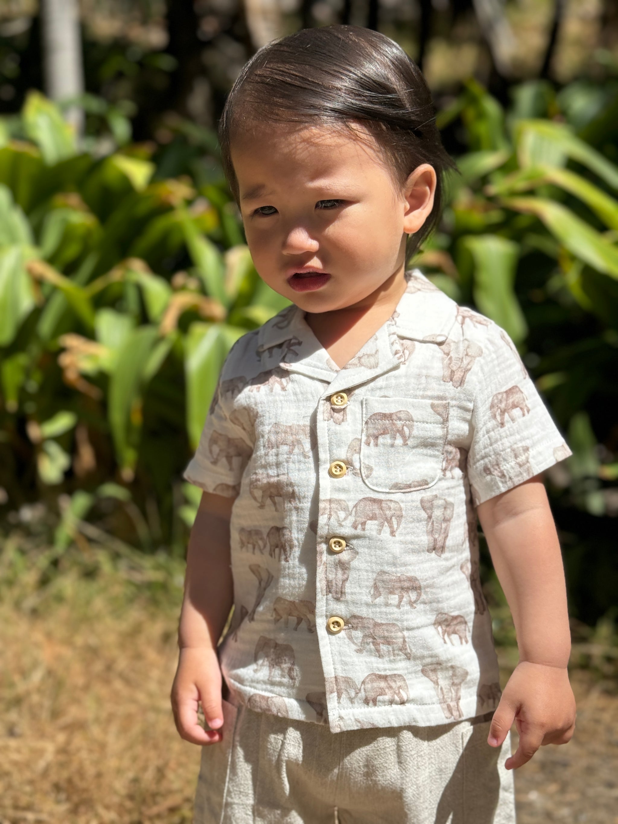 A toddler standing outdoors in front of green foliage, wearing a short-sleeve button-front shirt and matching pants in a light beige fabric with a brown elephant print.