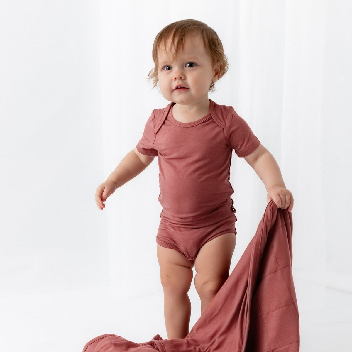 A toddler stands in a studio wearing a dusty rose short-sleeve bodysuit and holding a matching dusty rose sheet or blanket spread on the floor.