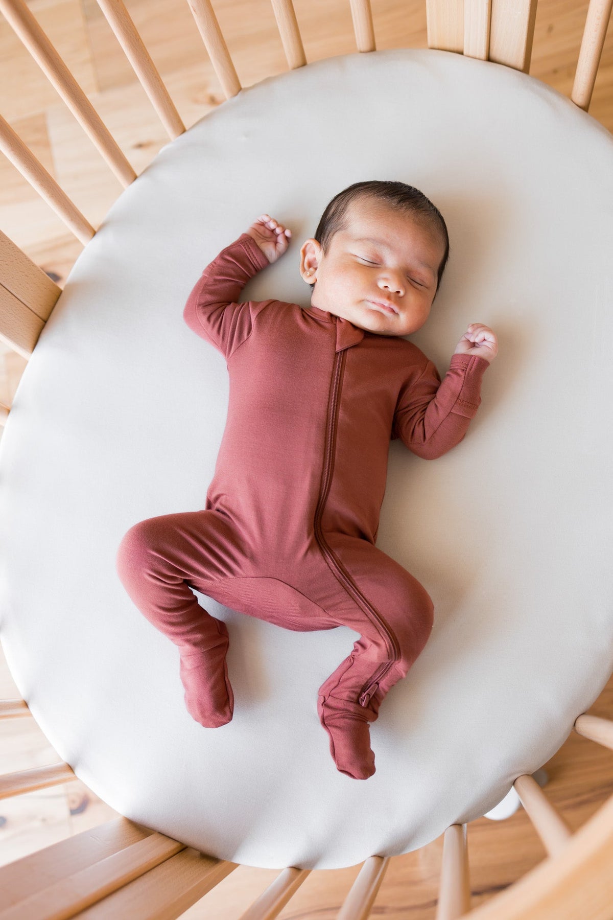 A sleeping newborn baby wearing a dusty rose footed zip-up sleeper, lying on a white oval mattress inside a wooden crib.