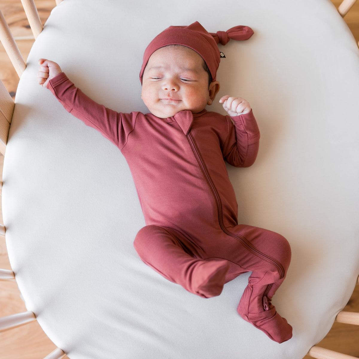 A newborn baby wearing a dusty rose terracotta long-sleeve zip-up sleeper and matching knotted beanie hat, lying on a white mattress in a crib.