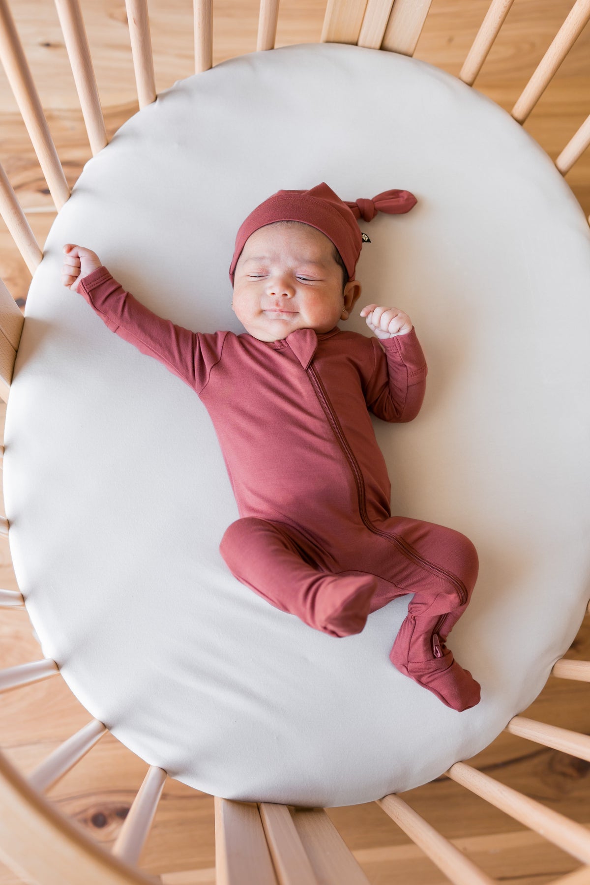 A newborn baby wearing a dusty rose terracotta long-sleeve zip-up sleeper and matching knotted beanie hat, lying on a white mattress in a crib.