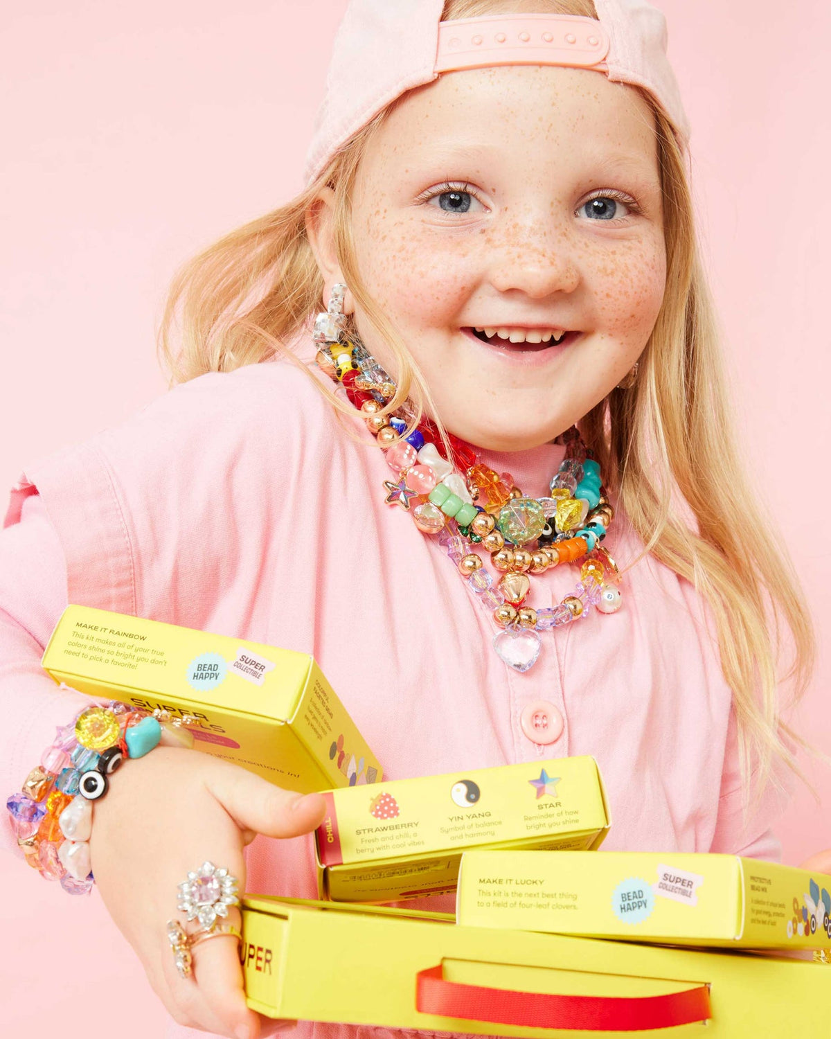 Child wearing colorful jewelry holding yellow boxes against a pink background