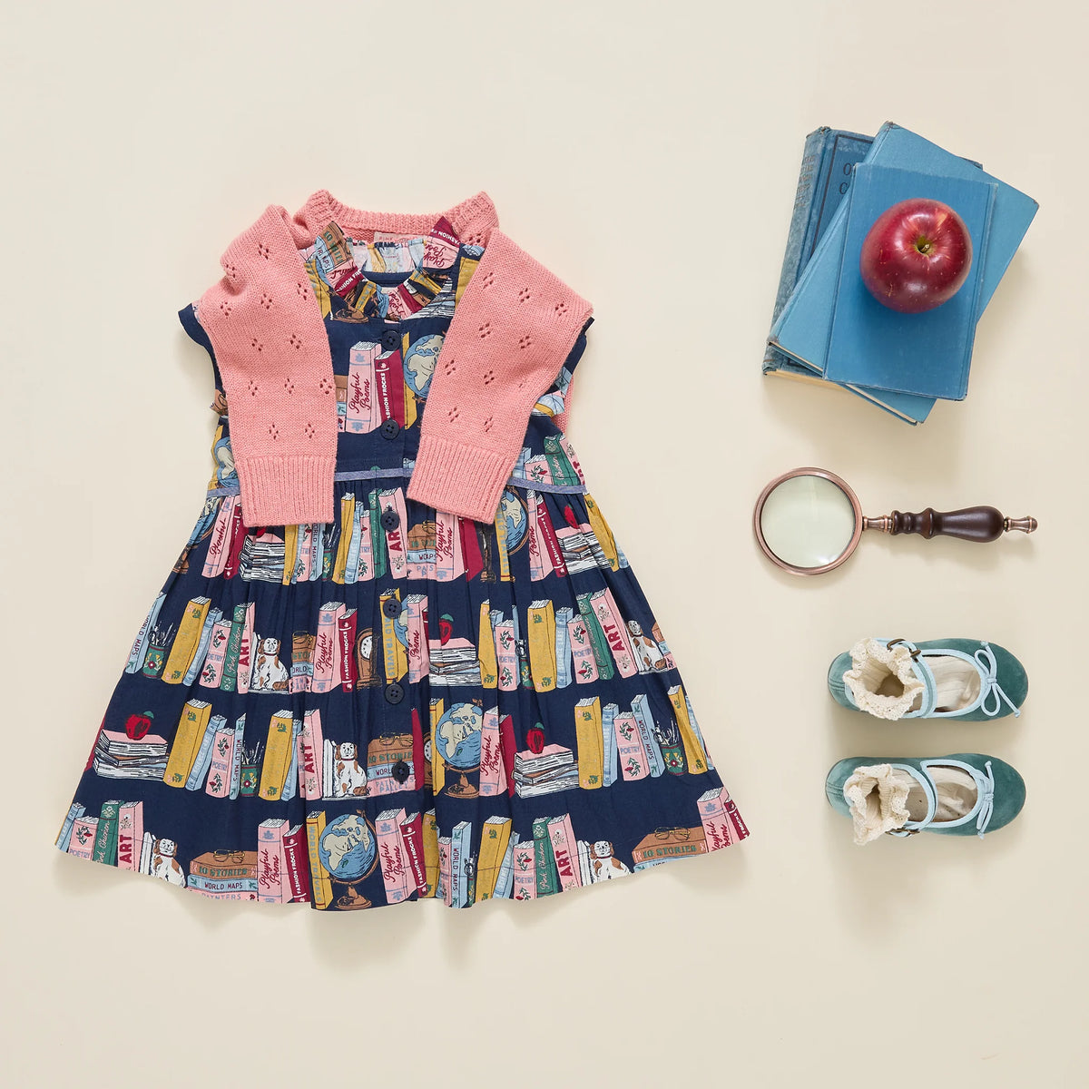 A flat-lay of a children's outfit set on a warm cream background, featuring a navy blue dress with a colorful book-print pattern and a pink knit cardigan. The outfit is styled with a stack of blue books and a red apple, a magnifying glass, and a pair of teal shoes.