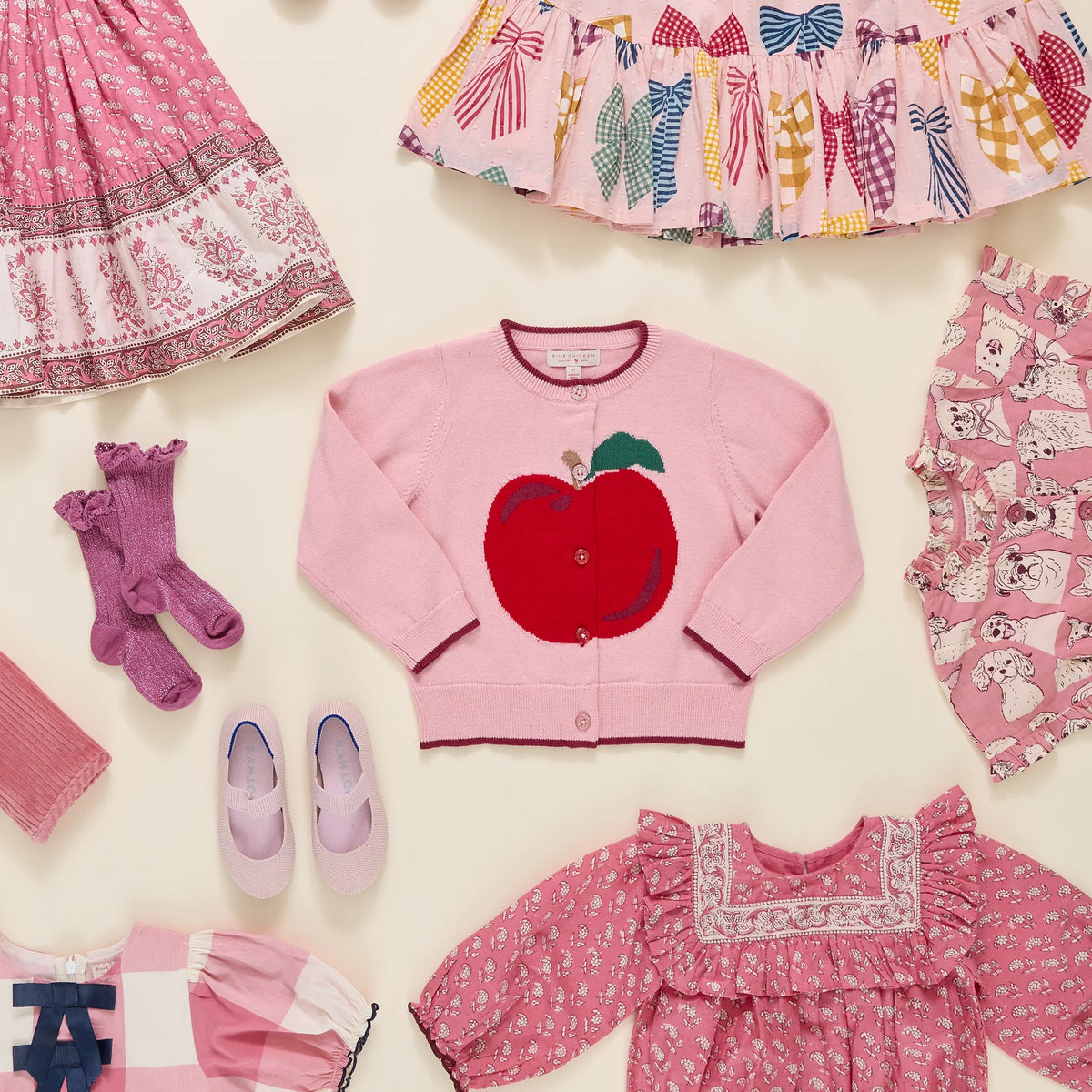A styled flat-lay of children's clothing on a light cream background, featuring a central pink cardigan with a large red apple applique, surrounded by matching pink patterned garments, socks, shoes, and a dress.