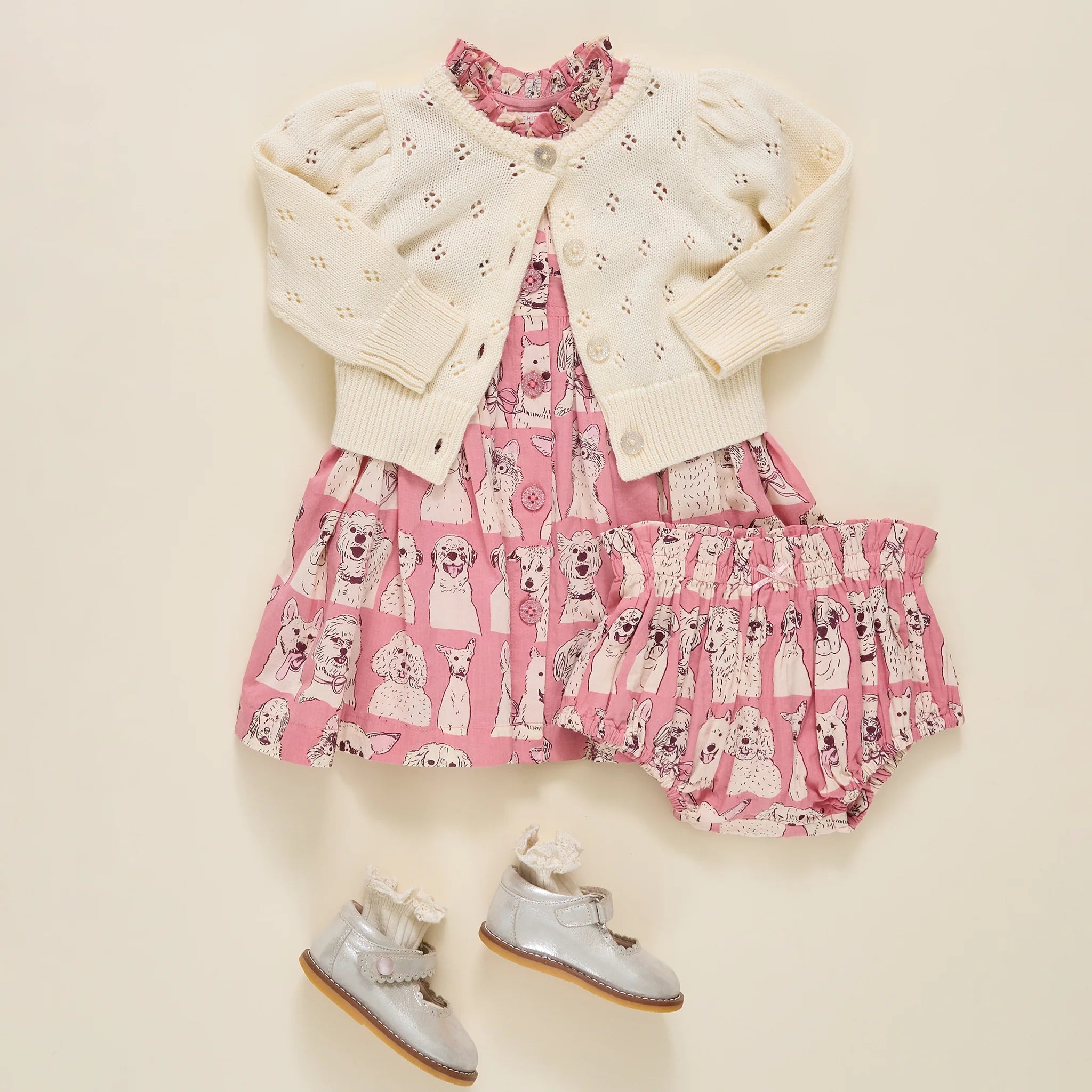 A baby girl's three-piece outfit set featuring a pink dress with a dog print, a cream eyelet cardigan, and a matching pink ruffled skirt, styled with a pair of silver baby shoes on a cream background.