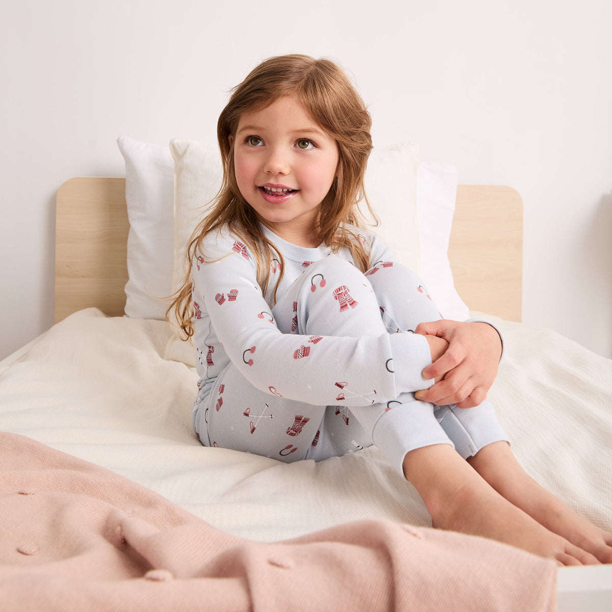 A young girl with light brown hair sits on a bed wearing a light gray long-sleeve pajama set with a red and pink pattern. The background features a light wood headboard, white pillows, and white bedding, with a soft pink blanket in the foreground.