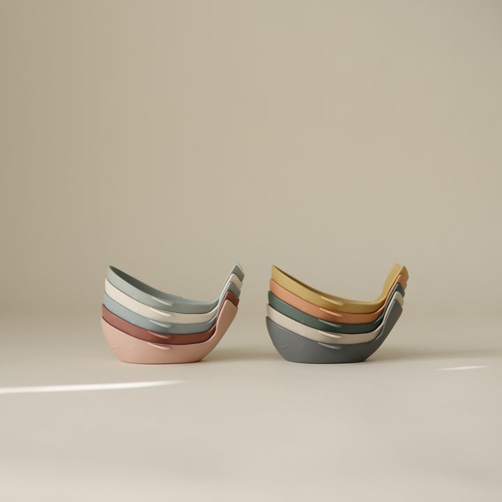 Two sets of stacked whale-shaped bath toys in muted, earthy tones resting on a neutral surface with soft lighting.
