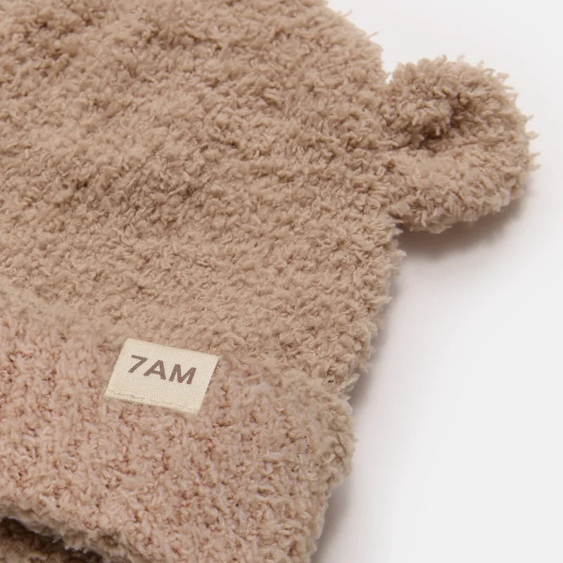 A close-up of a beige sherpa beanie with a small pom-pom on one side and a folded cuff featuring a cream-colored '7AM' brand label.