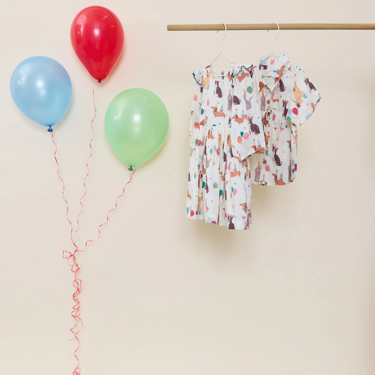 A baby dress and matching short-sleeve top with a colorful animal print hang on a wooden hanger bar against a beige wall, with three balloons (red, light blue, and light green) floating on the left.