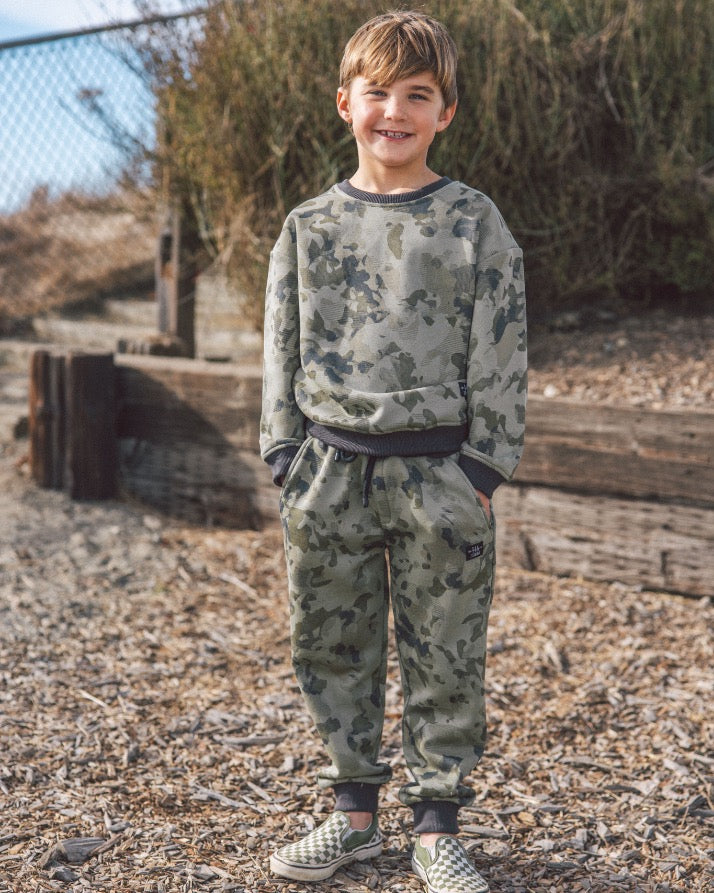 Child wearing a camouflage outfit standing outdoors near a wooden structure.
