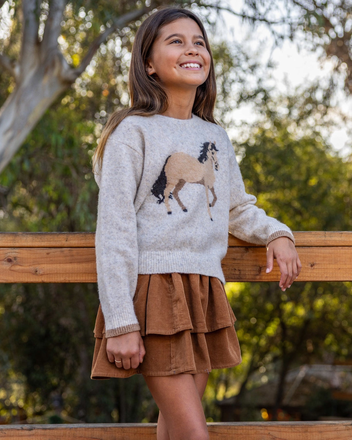 A young girl smiling and looking up while wearing a light gray knit sweater with a brown and beige horse design on the front, paired with a brown tiered skirt, leaning on a wooden fence outdoors.