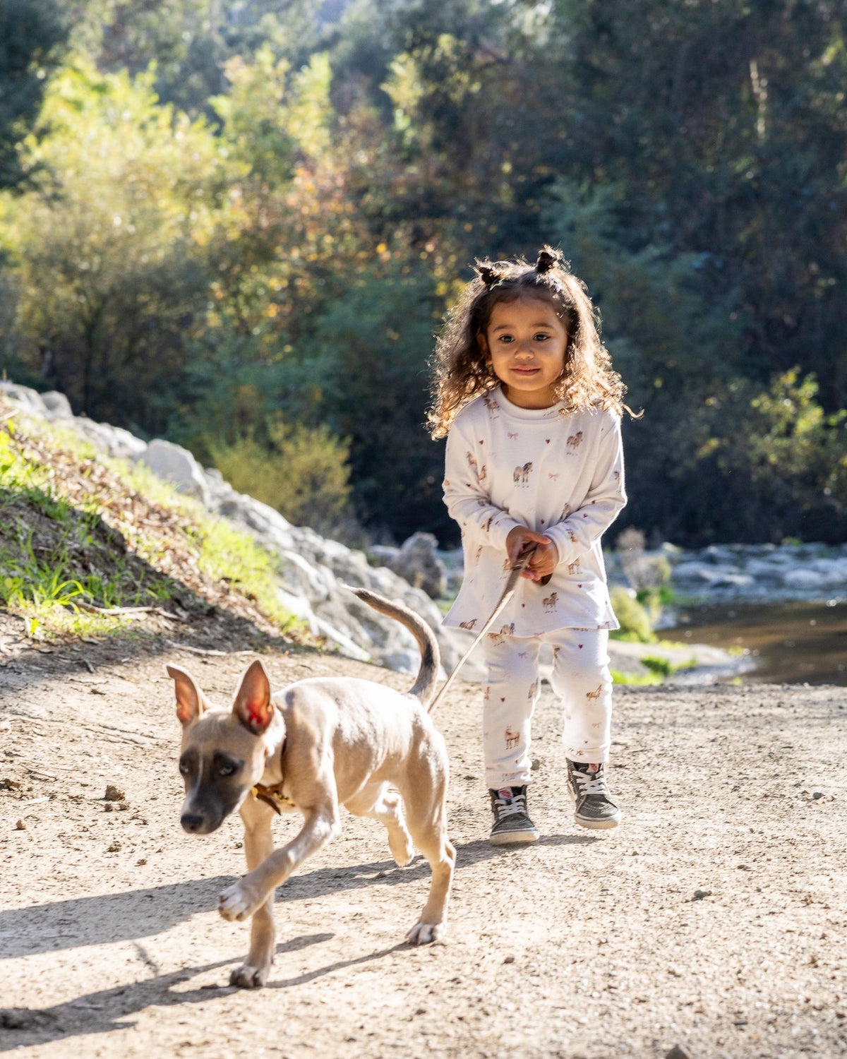 A young child wearing a matching light-colored pajama set walks outdoors on a dirt path with a small dog, with trees and rocks in the background.