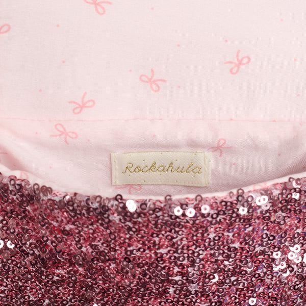 Close-up of a children's garment showing a light pink fabric with a subtle bow print, a central cream fabric label reading Rockahula, and a sequined hem in pink and burgundy sequins.