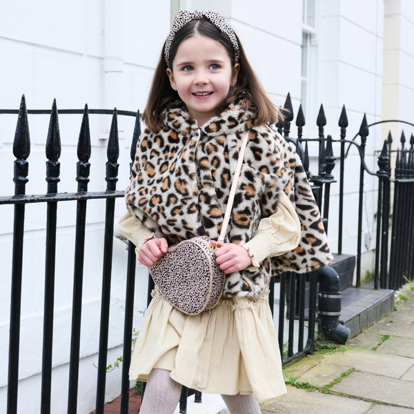 A young girl wearing a leopard-print faux fur hooded cape over a cream dress, holding a matching leopard-print crossbody bag and wearing a leopard-print headband, standing outdoors in front of a white building and black metal fence.