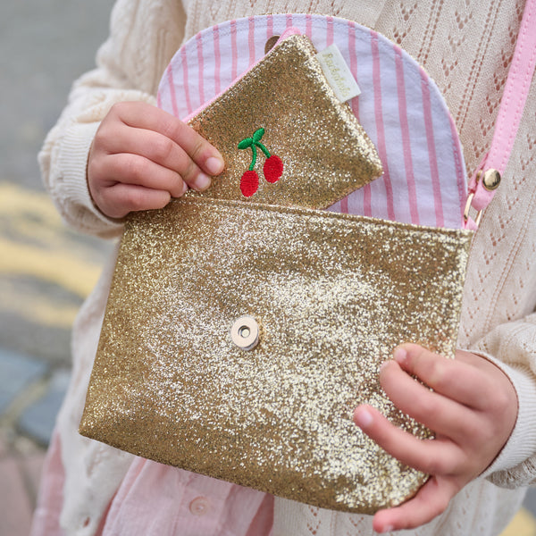 Child holding a glittery gold pouch with cherry design, wearing a pink and white checkered bag.