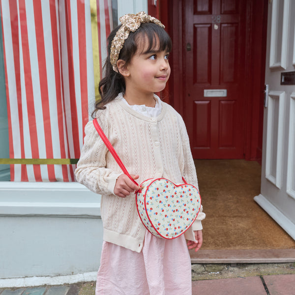A young girl wearing a cream cable-knit cardigan, pink skirt, and a gold floral headband carries a heart-shaped crossbody bag with a red cherry pattern and red strap, standing in front of a red door.